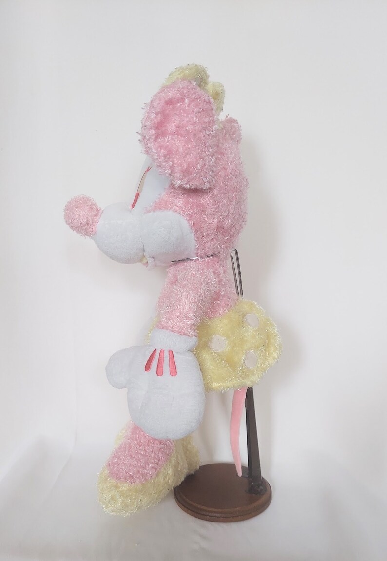 May include: A plush toy mouse in a side profile, featuring pink fur, white gloves with red accents, and a yellow polka-dotted skirt. The toy is mounted on a brown stand, showcasing its design and details.