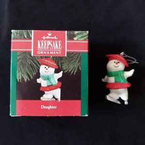 Peut inclure: Ornement Hallmark Keepsake représentant un bonhomme de neige. Le bonhomme de neige porte un chapeau et une jupe rouges, une écharpe verte et des patins à glace. L'ornement est à côté de sa boîte d'origine, qui indique "Daughter".