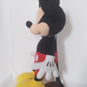 May include: A plush Mickey Mouse doll, with a black head and ears, a tan face, and a black body. The doll wears red shorts, white gloves, and yellow shoes. The doll is standing on a wooden base.