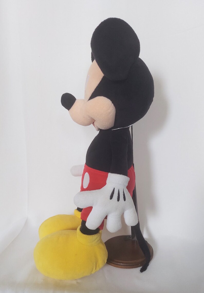 May include: A plush Mickey Mouse doll, with a black head and ears, a tan face, and a black body. The doll wears red shorts, white gloves, and yellow shoes. The doll is standing on a wooden base.