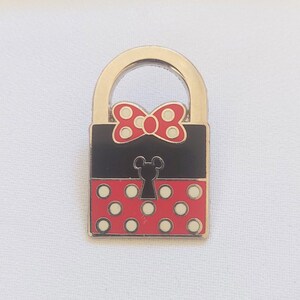 Puede incluir: Un candado dorado con un cuerpo rectangular negro y un diseño rojo con lunares blancos. El candado presenta un lazo rojo con lunares blancos y una cerradura con forma de cabeza de Mickey Mouse.