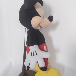May include: A plush Mickey Mouse toy, with a black head and ears, a tan face, white gloves, red shorts, yellow shoes, and a black body. The toy is displayed on a brown stand.