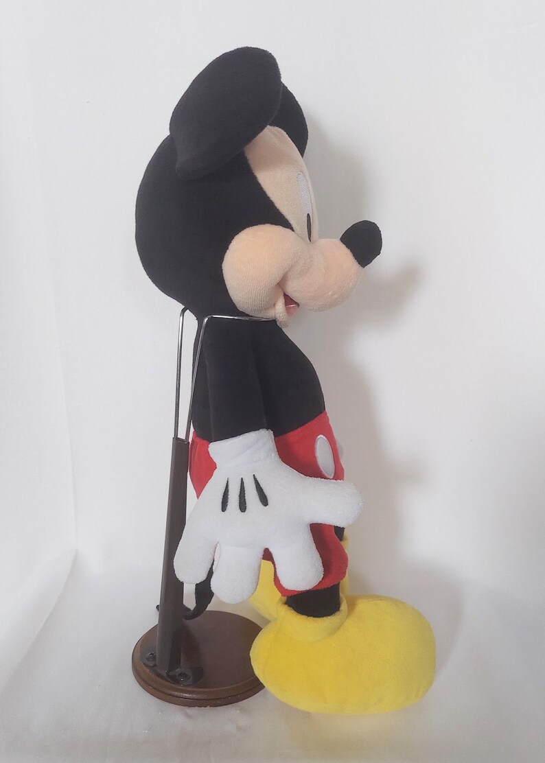 May include: A plush Mickey Mouse toy, with a black head and ears, a tan face, white gloves, red shorts, yellow shoes, and a black body. The toy is displayed on a brown stand.