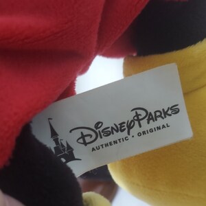 May include: A soft toy with red, black, and yellow sections. A white label shows the "Disney Parks AUTHENTIC ORIGINAL" logo and a castle image. The toy's design is inspired by a well-known cartoon character.