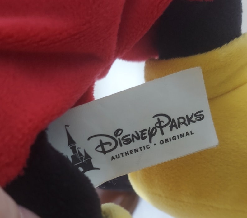 May include: A soft toy with red, black, and yellow sections. A white label shows the "Disney Parks AUTHENTIC ORIGINAL" logo and a castle image. The toy's design is inspired by a well-known cartoon character.