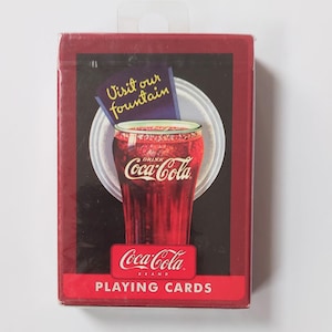 K&ouml;nnte beinhalten: Ein Kartenspiel der Marke Coca-Cola. Die Schachtel ist rot mit schwarzem Hintergrund und zeigt ein Glas Coca-Cola mit dem Text "Visit our fountain". Auf der Unterseite der Schachtel steht "Coca-Cola BRAND PLAYING CARDS".