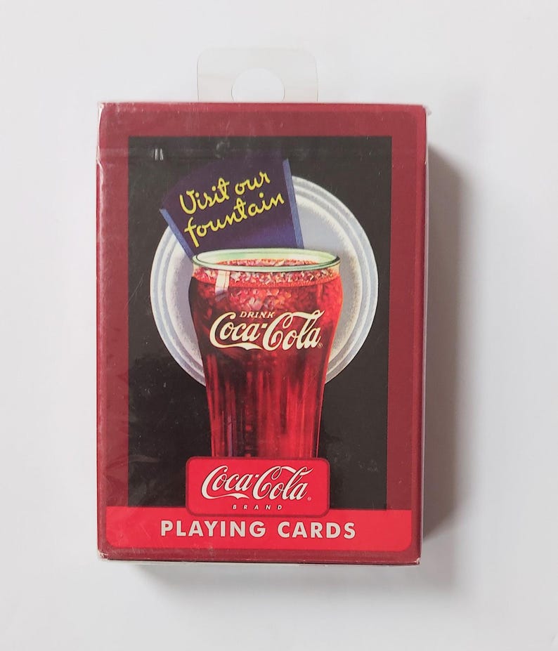 K&ouml;nnte beinhalten: Ein Kartenspiel der Marke Coca-Cola. Die Schachtel ist rot mit schwarzem Hintergrund und zeigt ein Glas Coca-Cola mit dem Text "Visit our fountain". Auf der Unterseite der Schachtel steht "Coca-Cola BRAND PLAYING CARDS".