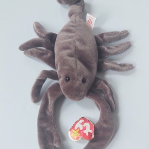 Peut inclure: Un jouet scorpion en peluche marron avec une texture douce semblable à du velours, doté de huit pattes, deux grandes pinces et une queue courbée. Une étiquette Ty Beanie Baby rouge et jaune en forme de cœur est fixée sur le côté.