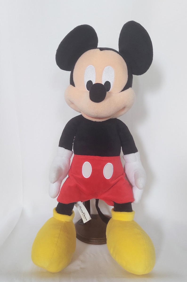 May include: A plush Mickey Mouse doll, with a black torso and ears, red shorts, white gloves, and yellow shoes. The doll has a cream-colored face with a black nose and white eyes. The Disney Parks tag is visible.