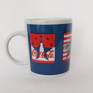 Puede incluir: Taza de cerámica blanca con fondo azul con estrellas y rayas rojas, y el logotipo de Coca-Cola. La taza tiene un asa blanca y un interior blanco.