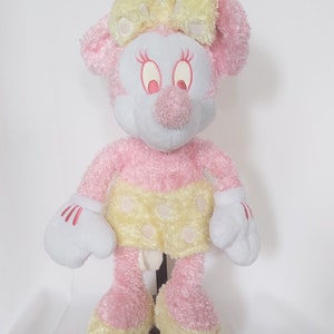 May include: A plush Minnie Mouse doll in pink, white, and yellow. The doll has a pink body, a white face, and a yellow bow. She wears a yellow skirt with white polka dots and white gloves with red accents.