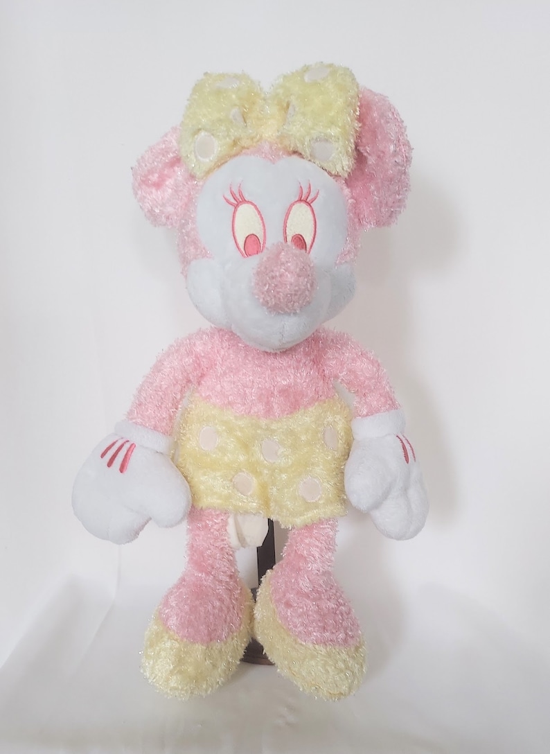May include: A plush Minnie Mouse doll in pink, white, and yellow. The doll has a pink body, a white face, and a yellow bow. She wears a yellow skirt with white polka dots and white gloves with red accents.