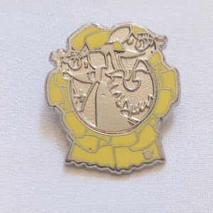May include: A silver-colored pin with a yellow border and a Disney character design. The pin features a central image of a character with a yellow hat and a yellow banner. The Disney logo is visible on the bottom right.