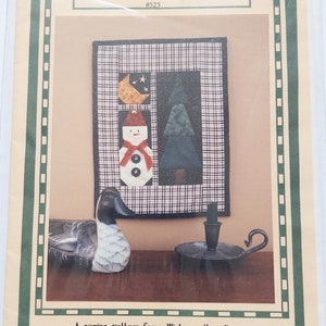 Op de afbeelding: Een naaipatroon genaamd "Winter Nights" van Sweet Peas, Inc. voor een winterwandkleed. Het ontwerp toont een sneeuwpop, een boom en een maan. Een decoratieve eend en een kandelaar zijn ook zichtbaar.