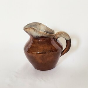 May include: A small ceramic pitcher with a glossy dark brown body and a curved handle. The rim and the top of the handle feature a contrasting light beige and cream-colored glaze that drips down slightly over the brown surface of the vessel.
