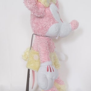 May include: A plush toy of a cartoon mouse, primarily pink and yellow, with white gloves and a red bow. The toy is standing on a metal stand with a wooden base. The mouse has a cheerful expression.