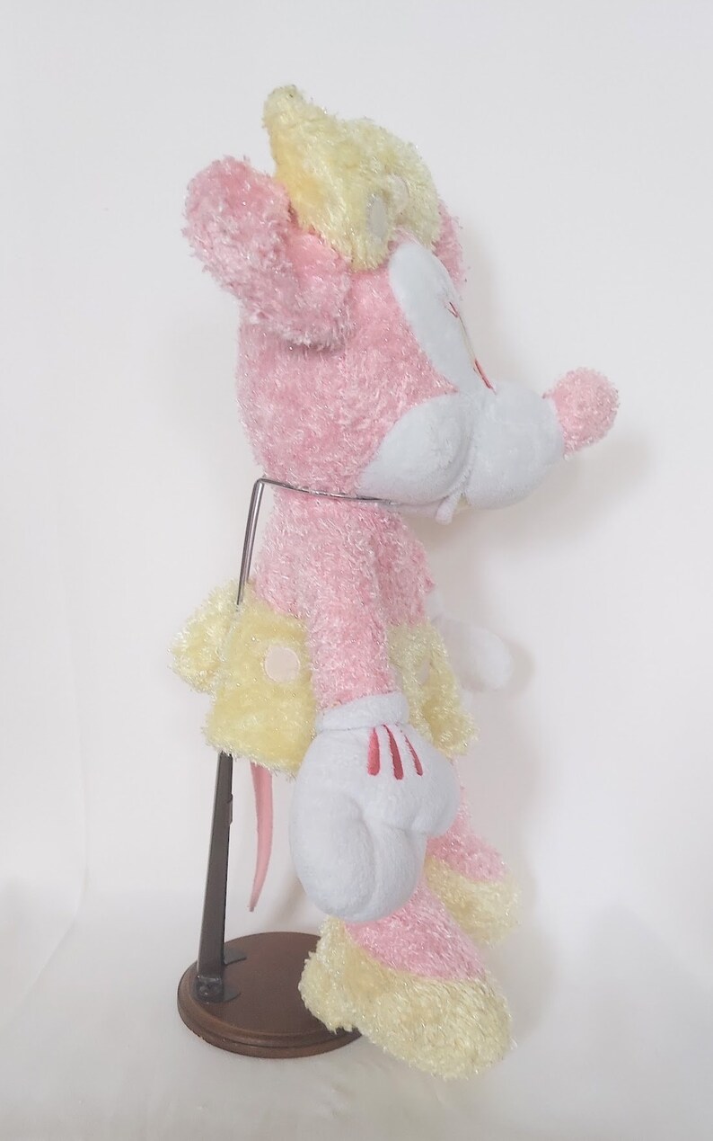 May include: A plush toy of a cartoon mouse, primarily pink and yellow, with white gloves and a red bow. The toy is standing on a metal stand with a wooden base. The mouse has a cheerful expression.