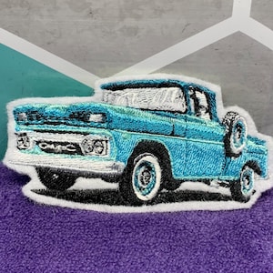 May include: Embroidered patch of a vintage teal GMC pickup truck with white accents. The truck has a spare tire on the side and is set against a white border. The GMC logo is visible on the front.