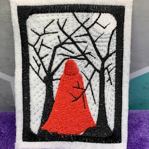 May include: Embroidered patch featuring a red hooded figure in a forest scene. The design is framed by a black border on a white background. The trees are black, and the figure is in a red cloak. The patch is rectangular.