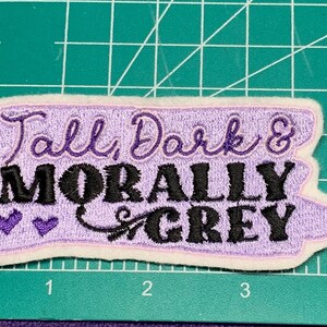 May include: Embroidered patch with the text "Tall, Dark & Morally Grey" in purple and black. The patch has a light purple background with a white border and two small purple hearts.