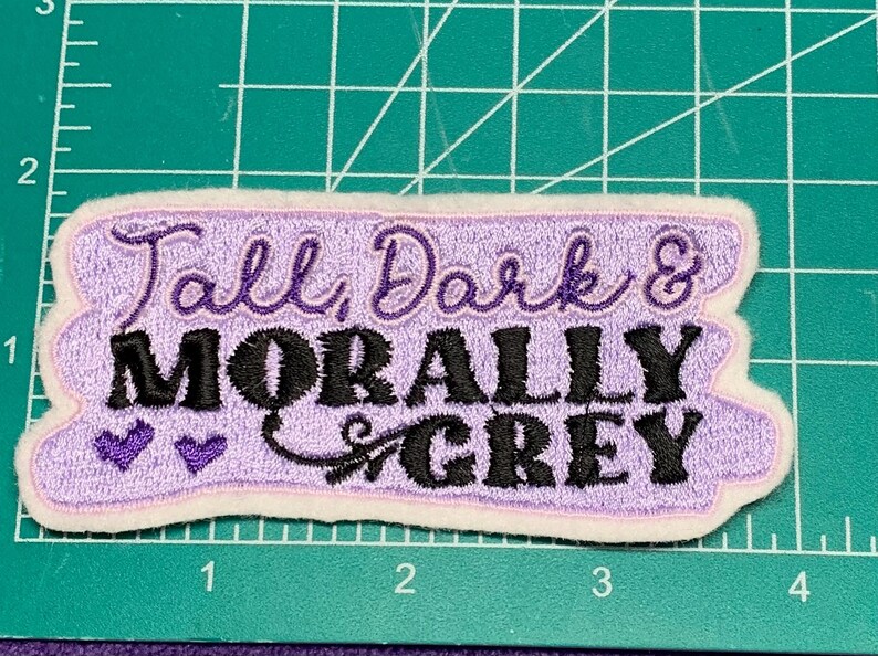 May include: Embroidered patch with the text "Tall, Dark & Morally Grey" in purple and black. The patch has a light purple background with a white border and two small purple hearts.