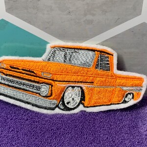 May include: Embroidered patch of an orange vintage pickup truck with silver and black detailing. The truck has a white border and is set against a purple background.