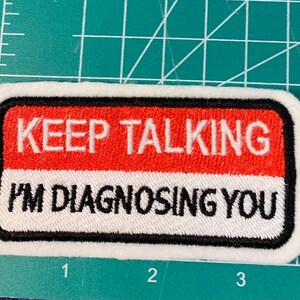 Puede incluir: Parche blanco bordado con borde negro. El parche tiene texto rojo y blanco que dice "KEEP TALKING I'M DIAGNOSING YOU".