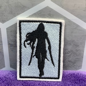 May include: A white embroidered patch with a black silhouette of a person holding two swords. The background of the patch is light blue.