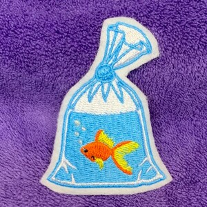 May include: An embroidered patch featuring a clear plastic bag filled with blue water and a small orange goldfish with yellow fins. The bag is tied at the top with blue thread and includes white embroidered bubbles rising from the fish.