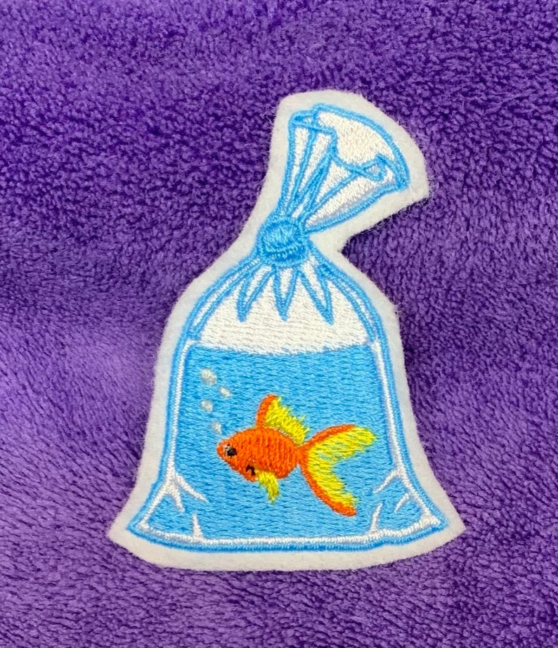 May include: An embroidered patch featuring a clear plastic bag filled with blue water and a small orange goldfish with yellow fins. The bag is tied at the top with blue thread and includes white embroidered bubbles rising from the fish.