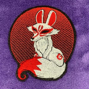 May include: Embroidered patch featuring a white fox with red accents, including the ears, tail, and facial markings. The fox is set against a red circular background with a black border, all on a purple fabric.