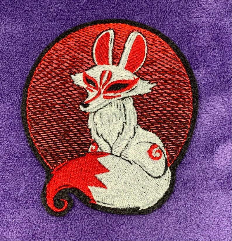 May include: Embroidered patch featuring a white fox with red accents, including the ears, tail, and facial markings. The fox is set against a red circular background with a black border, all on a purple fabric.