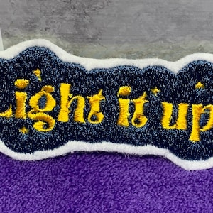 May include: A blue and white embroidered patch with the text "Light it up" in yellow. The patch has a cloud-like shape and is decorated with small yellow stars.