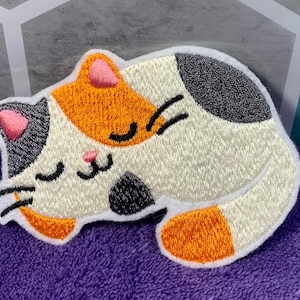 Op de afbeelding: Een geborduurde patch van een slapende kat, in de kleuren wit, oranje en grijs. De kat ligt opgerold met zijn pootjes onder zijn kin.
