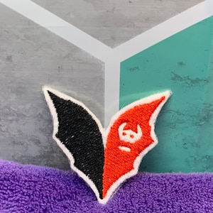 May include: A black and red embroidered patch in the shape of a bat with a white face. The patch is on a purple fuzzy surface.