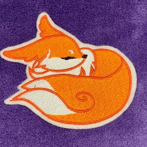 May include: Embroidered patch featuring a sleeping fox. The fox is orange with white accents and a black eye, outlined in white. The patch is on a purple background. The fox is curled up, with its tail wrapped around its body.