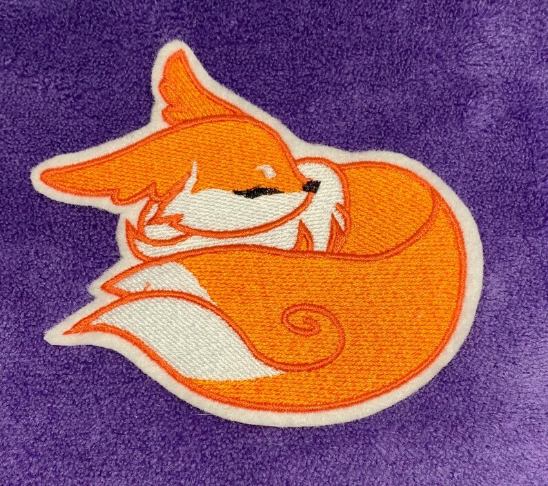 May include: Embroidered patch featuring a sleeping fox. The fox is orange with white accents and a black eye, outlined in white. The patch is on a purple background. The fox is curled up, with its tail wrapped around its body.