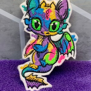 May include: A colorful embroidered patch of a cartoon dragon with large green eyes. The dragon is purple, blue, yellow, and green with a white background.