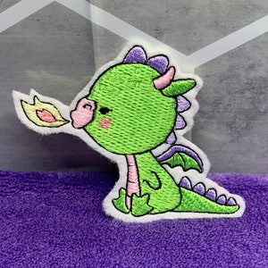 May include: Green embroidered dragon applique with pink cheeks, purple horns, and a yellow flame coming out of its mouth. The dragon is sitting on a white background.