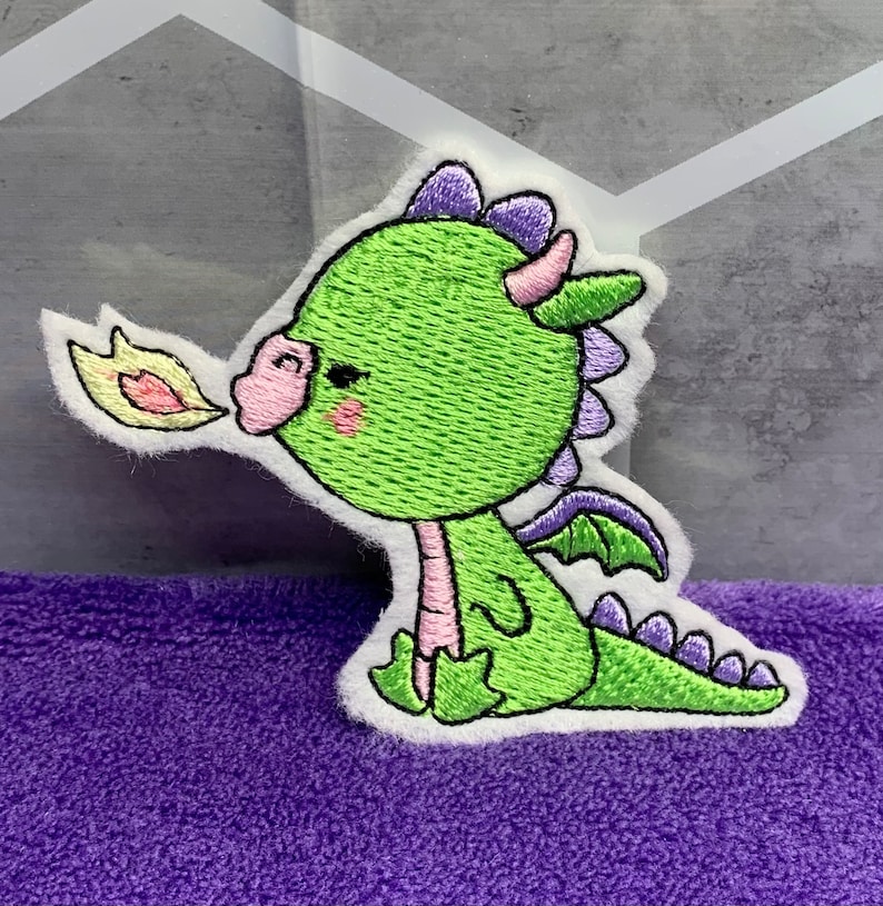 May include: Green embroidered dragon applique with pink cheeks, purple horns, and a yellow flame coming out of its mouth. The dragon is sitting on a white background.