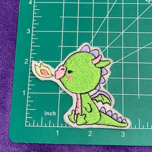 May include: Green embroidered iron-on patch of a cartoon dragon with purple accents blowing fire. The dragon has a pink nose and closed eyes.