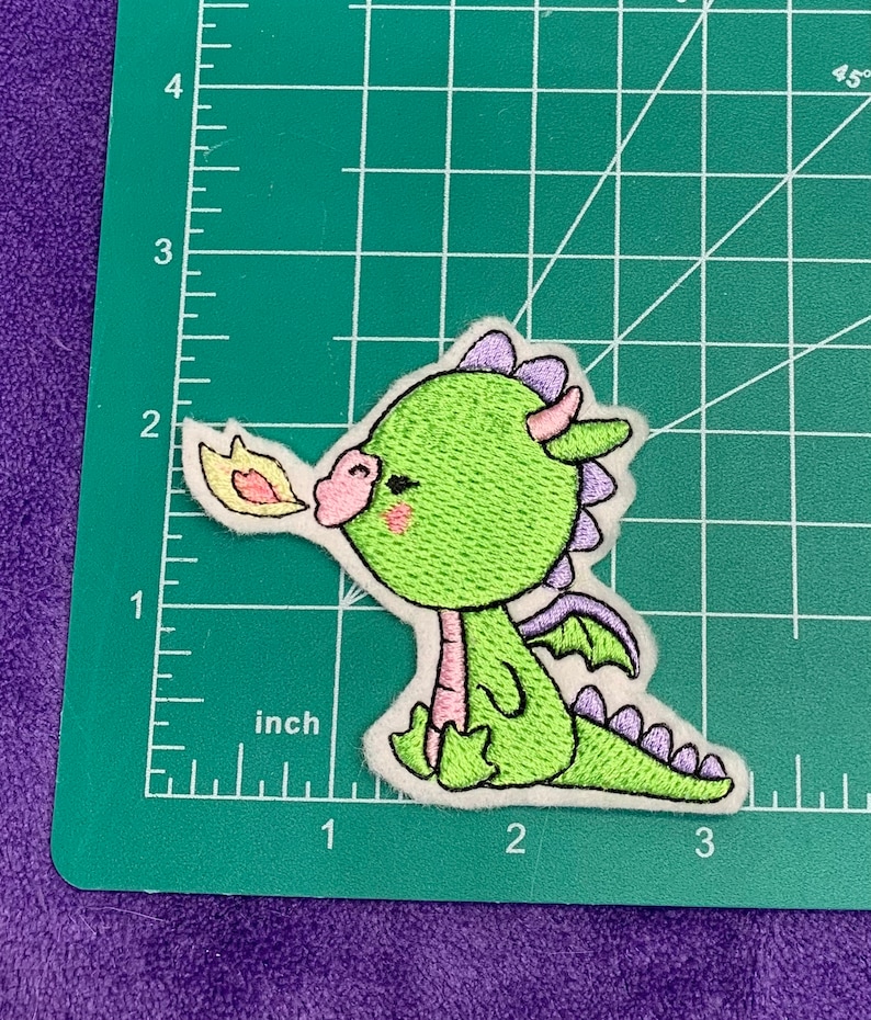 May include: Green embroidered iron-on patch of a cartoon dragon with purple accents blowing fire. The dragon has a pink nose and closed eyes.