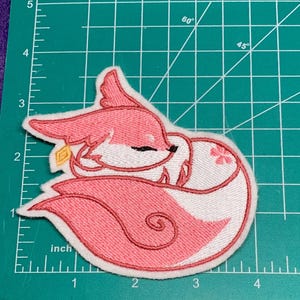 May include: Embroidered patch of a pink fox curled up. The fox has white and pink fur, with a small gold charm. The patch is approximately 7.6 cm wide.