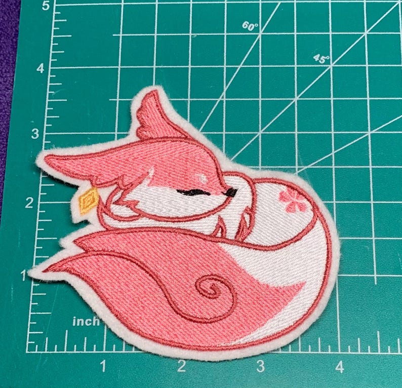 May include: Embroidered patch of a pink fox curled up. The fox has white and pink fur, with a small gold charm. The patch is approximately 7.6 cm wide.