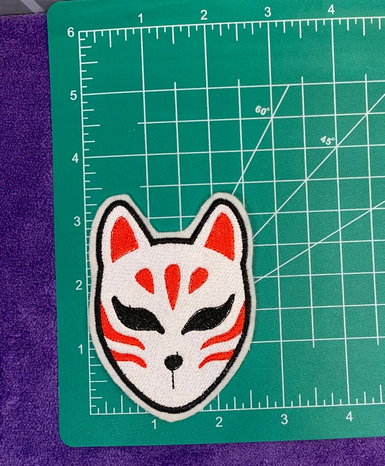 May include: White embroidered fox mask with red and black details. The mask is on a green cutting mat with a ruler.