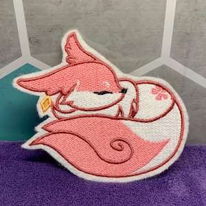 May include: Embroidered patch featuring a sleeping fox in shades of pink and white. The fox is curled up, with a small gold-colored charm near its ear. The patch has a white border and is set against a purple surface.
