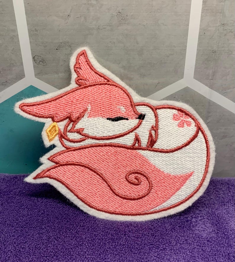 May include: Embroidered patch of a sleeping fox in pink and white. The fox is curled up, with a small gold charm near its ear. The patch has a white border and is set against a purple surface.