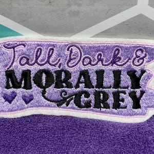 May include: Embroidered patch with the text "Tall, Dark & Morally Grey" in black and purple. The patch has a white border and a purple background. Two small purple hearts are below the word "Morally".