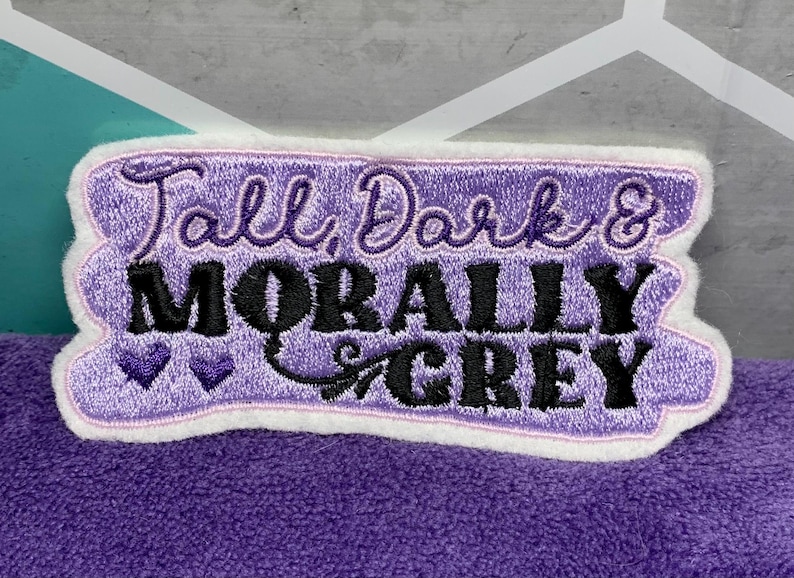 May include: Embroidered patch with the text "Tall, Dark & Morally Grey" in black and purple. The patch has a white border and a purple background. Two small purple hearts are below the word "Morally".