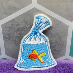 May include: An embroidered patch featuring a goldfish inside a clear plastic bag filled with water. The fish is orange with yellow fins, and the bag is tied at the top with white thread. The design includes small white bubbles in the blue water.